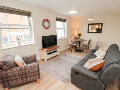 A living room with a grey sofa and a plaid armchair a TV on a white stand and a round dining table with chairs at Sunny Bay in The Bay near Filey
