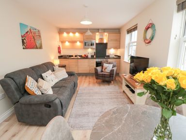 A living room with a gray sofa and kitchen area with wooden cabinets and a window with blinds at Sunny Bay The Bay near Filey