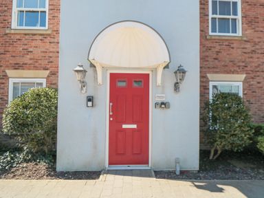 A red front door with an arched white canopy and two wall lanterns flanked by bushes at Sunrise in The Bay near Filey