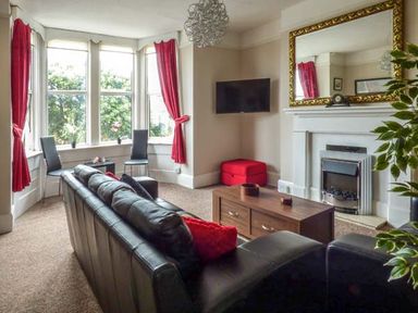 A living room with a black leather sofa red curtains a wooden coffee table a wall-mounted TV and a fireplace at Westgate Retreat in Hunstanton