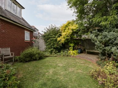 A garden with a lawn surrounded by bushes and trees with wooden benches near a red brick house at Hurst Green in Ewyas Harold near Hereford
