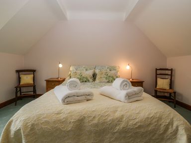 A bedroom with a bed covered in a patterned yellow bedspread with two rolled white towels on top two wooden nightstands with lamps and two wooden chairs with cushions at Hurst Green Ewyas Harold near Hereford