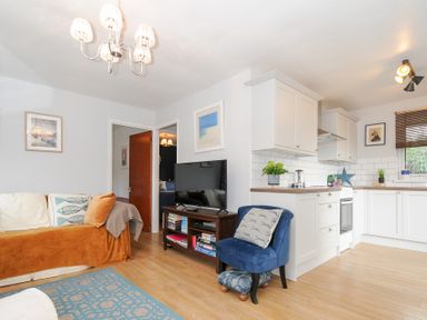 A living room with a sofa a blue chair and a TV next to a kitchen with white cabinets at No 1 The Heathers Rosecraddoc Lodge near Liskeard