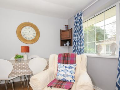 A dining area with a polka dot tablecloth on a table surrounded by white chairs and a cream armchair with a fish-pattern pillow near a window with blue and white curtains at No 1 The Heathers Rosecraddoc Lodge near Liskeard