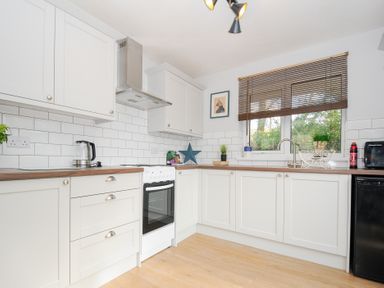 A kitchen with white cabinets a stove a kettle and a window with blinds at No 1 The Heathers Rosecraddoc Lodge near Liskeard