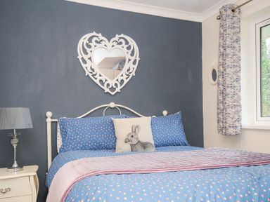 A bedroom with a bed with blue and pink bedding and a heart-shaped mirror on a dark wall at No 1 The Heathers Rosecraddoc Lodge near Liskeard
