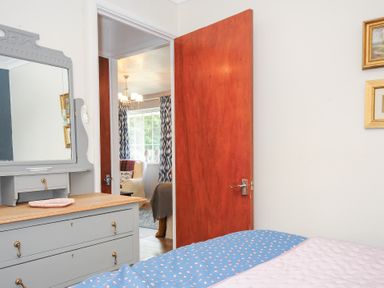 A bedroom with a blue and pink bedspread a gray dresser with a mirror and an open wooden door showing a view into a living room at No 1 The Heathers Rosecraddoc Lodge near Liskeard