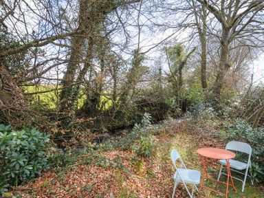 A small outdoor seating area with two white chairs and an orange table near trees and shrubs at No 1 The Heathers Rosecraddoc Lodge near Liskeard