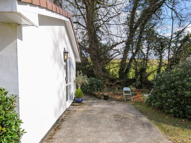 A white house exterior with a doorway and outdoor seating on a concrete patio surrounded by trees at No 1 The Heathers Rosecraddoc Lodge near Liskeard