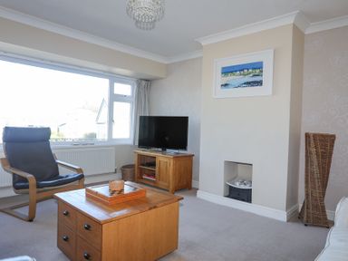A living room with a wooden coffee table a wooden TV stand with a television a black armchair a small fireplace and a wicker basket at Tegfan in Trearddur Bay