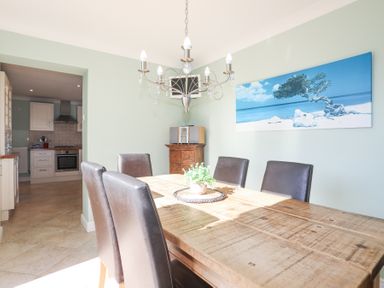 A dining room with a wooden table and six black chairs a chandelier a painting of a beach and tree on the wall and a small wooden cabinet with a stereo system at Tegfan in Trearddur Bay