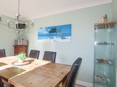 A dining room with a wooden table surrounded by black chairs a glass cabinet with decorative items and a landscape painting on the wall at Tegfan in Trearddur Bay