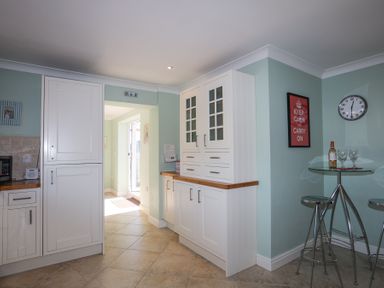 A kitchen with white cabinets, a small glass table with two metal stools, a clock, and a framed poster at Tegfan in Trearddur Bay
