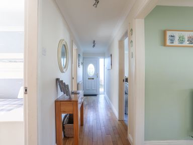 A hallway with a wooden console table and round mirror leading to a white front door at Tegfan in Trearddur Bay