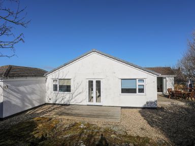 The exterior view of a single-story white house with a wooden deck and gravel yard at Tegfan in Trearddur Bay