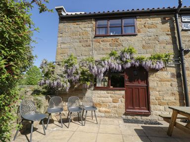 An outdoor patio with four chairs and a wooden picnic table next to a stone building with a wooden door and windows with flowering plants above at Goathland Cottage in Ruswarp near Whitby