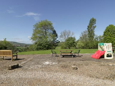 An outdoor area with a wooden bench, two metal chairs, a picnic table, and a plastic children's slide surrounded by trees and grass at Goathland Cottage in Ruswarp near Whitby