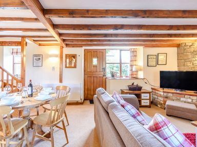 A living and dining area with a wooden dining table set with white dishes a beige sofa with plaid pillows a wooden door and a flat screen TV at Goathland Cottage Ruswarp near Whitby