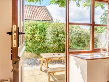 A wooden door opening to a patio with a wooden picnic table and green bushes at Goathland Cottage in Ruswarp near Whitby