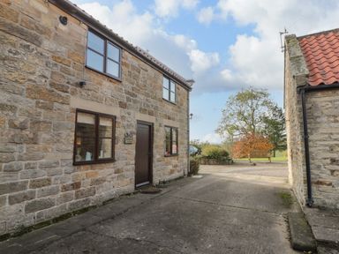 Exterior of a stone cottage with windows and a brown door next to a driveway and garden bench at Goathland Cottage in Ruswarp near Whitby