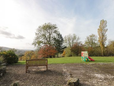 An outdoor area with a wooden bench on gravel and a children's play slide on grass surrounded by trees at Goathland Cottage in Ruswarp near Whitby