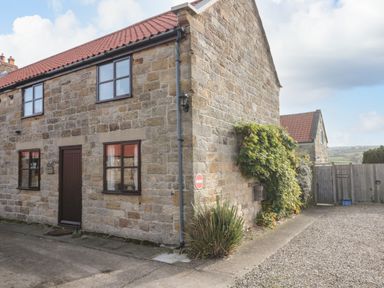 A stone cottage with dark framed windows and a brown door beside a gravel driveway and a wooden fence at Goathland Cottage in Ruswarp near Whitby
