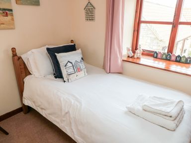 A single bed with white bedding and decorative pillows next to a window with pink curtains and ornaments at Goathland Cottage Ruswarp near Whitby
