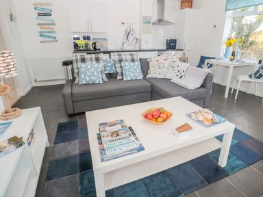 A living room with a gray sofa and blue patterned cushions a white coffee table with magazines fruit and seashells a blue rug and a white dining table with chairs by a window at Bracken Barn Cottage in Widdrington near Amble