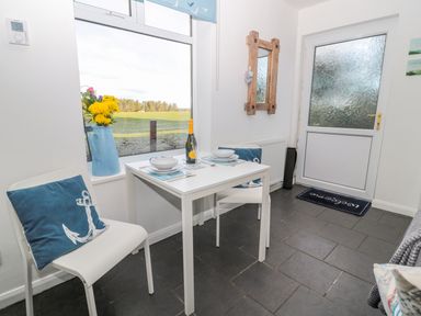 A dining area with a small white table set for two near a window with a blue pitcher of flowers and navy blue cushions on white chairs at Bracken Barn Cottage in Widdrington near Amble