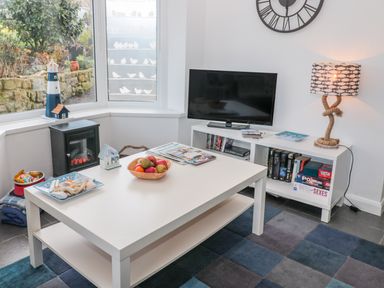 A living room with a white coffee table holding fruit and magazines a television on a white stand with books and board games and a lamp with a rope base at Bracken Barn Cottage in Widdrington near Amble