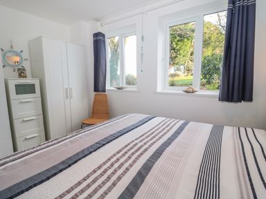 A bedroom with a bed striped bedding a white wardrobe a tall white cabinet a wicker chair and two windows with a view of trees at Bracken Barn Cottage in Widdrington near Amble