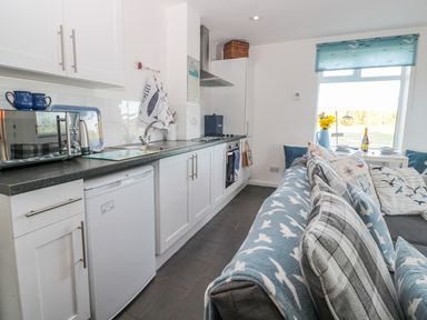 A kitchen with white cabinets a microwave and a small refrigerator next to a living area with a sofa and cushions at Bracken Barn Cottage in Widdrington near Amble