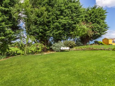 A grassy garden with trees a bench with pillows and a wooden shed at Bracken Barn Cottage in Widdrington near Amble
