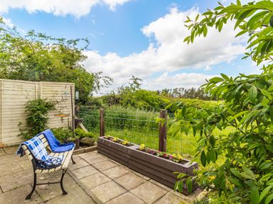 A garden patio with a bench covered by blankets and a rectangular flower planter with a wire fence and greenery in the background at Bracken Barn Cottage in Widdrington near Amble