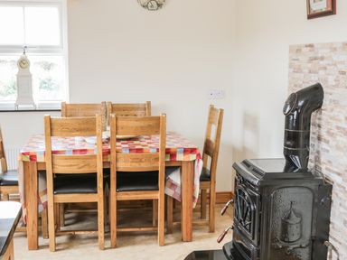 A dining area with a wooden table covered by a checkered tablecloth and six wooden chairs next to a black stove with a pipe in a room with a window and a clock at McGuire's Cottage Barnatra County Mayo