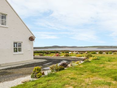An outdoor view with part of a white house a gravel driveway and a garden overlooking a body of water and hills at McGuire's Cottage in Barnatra County Mayo