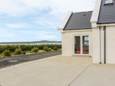 The exterior of a cream house with slate roof and glass double doors opening to a concrete driveway and garden with shrubbery at McGuire's Cottage Barnatra County Mayo