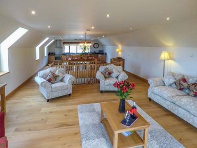 An open living room with patterned armchairs and sofa a coffee table with flowers and wine and a dining area with wooden table and chairs at Brynhir Farm in Howey near Llandrindod Wells