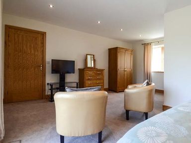 A room with two beige chairs a wooden door a small television on a stand a wooden chest of drawers with a mirror and a wooden wardrobe at Brynhir Farm in Howey near Llandrindod Wells