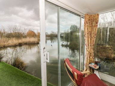 A red chair and small table by a large glass door overlooking a lake with reeds and trees at Secret Island Yurt in Beckford near Tewkesbury
