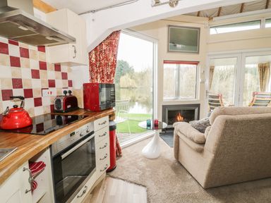 A kitchen with a wooden countertop, red kettle, toaster, microwave, and oven next to a living area with a beige sofa, small round white table, fireplace, and glass doors showing an outside view at Secret Island Yurt in Beckford near Tewkesbury