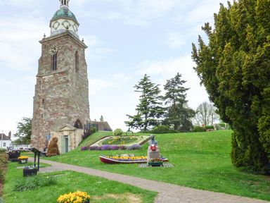 A stone clock tower in a park with flower beds a statue and trees at Secret Island Yurt in Beckford near Tewkesbury