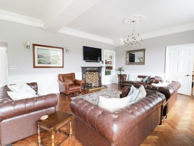 A living room with brown leather sofas a brown armchair a wall-mounted television a fireplace with stacked firewood a rug a chandelier and framed artwork at Bryn Derwen in Bethesda near Caernarfon