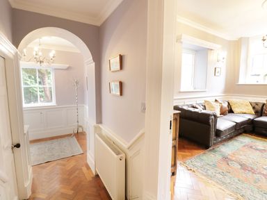 An interior hallway with a chandelier and coat rack next to a living room with a leather sofa and patterned rug at Bryn Derwen Bethesda near Caernarfon