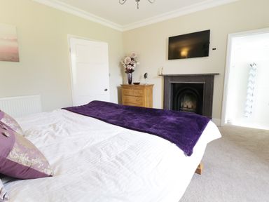 A bedroom with a bed a purple throw pillows a fireplace a wooden dresser a television and a view into a bathroom at Bryn Derwen Bethesda near Caernarfon