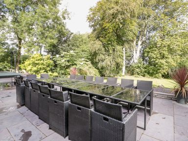 An outdoor patio with a long glass dining table and multiple black wicker chairs surrounded by trees and plants at Bryn Derwen in Bethesda near Caernarfon