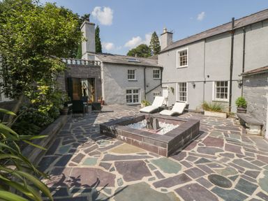 An outdoor patio area with stone flooring a square fire pit two lounge chairs a table with chairs and surrounding plants at Bryn Derwen Bethesda near Caernarfon