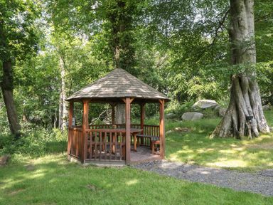 A wooden gazebo with a table and benches in a grassy wooded area at Bryn Derwen in Bethesda near Caernarfon