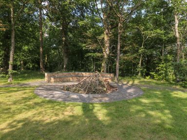 A circular stone fire pit with stacked branches and a curved brick bench in a grassy wooded area at Bryn Derwen Bethesda near Caernarfon