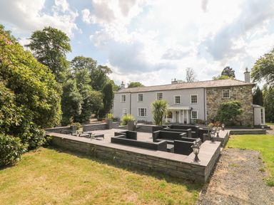 A stone patio with outdoor furniture and plants in front of a large house with white and stone walls at Bryn Derwen in Bethesda near Caernarfon